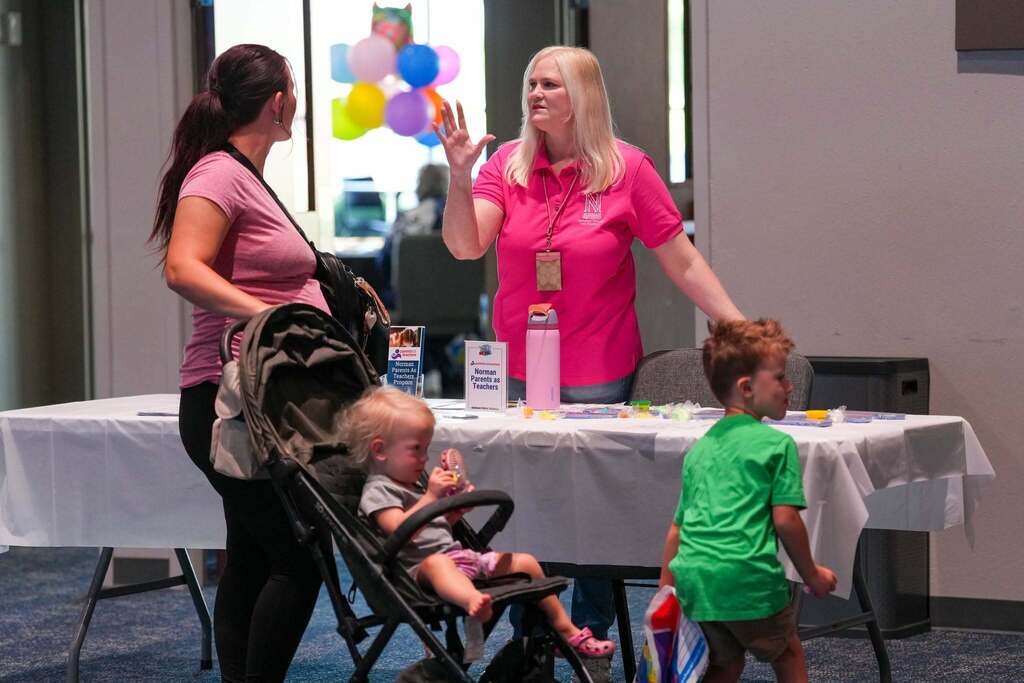Two women and two children are near a table with balloons in a room with a window.