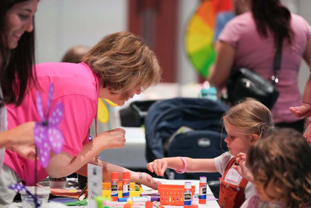 A woman assists a child with crafts at a table; other children and adults are nearby.