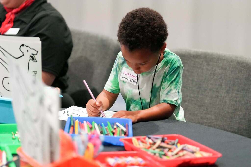 A child uses a pen to color a sheet of paper. Various colored pencils and markers are on the table.