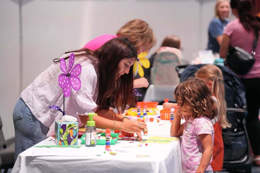 A woman and a girl work at a table with craft materials. Other people, including one with a stroller, are in the background.
