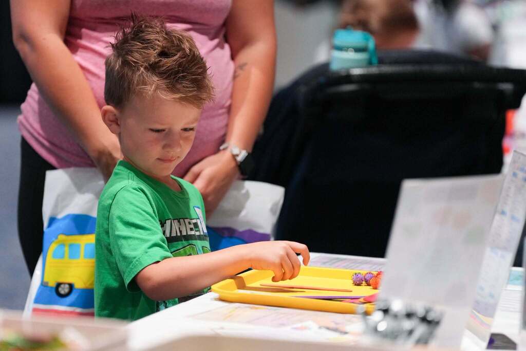 A child in green works on a tray with food items, seated at a table with an adult nearby.