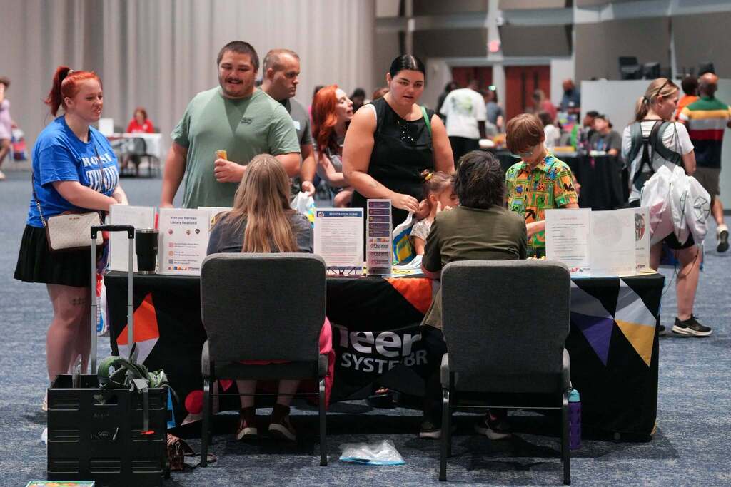 A group of people at a convention stand, with some seated and others standing, looking at items on tables.