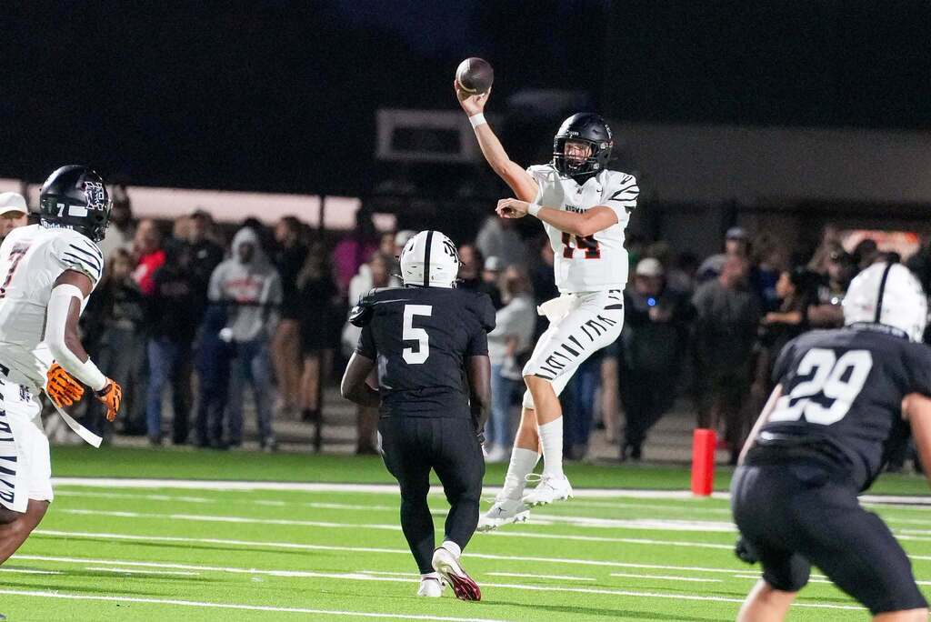 A football player in white jumps and throws a ball. Two opponents in black. Spectators in the background.