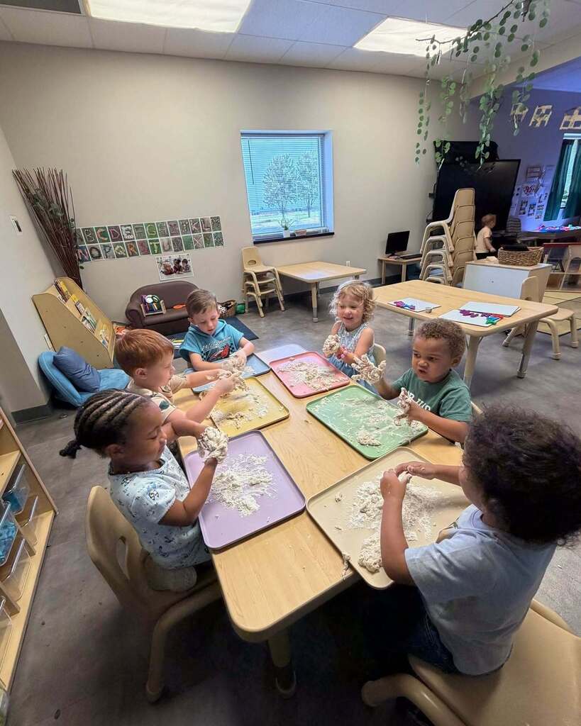 A classroom with children seated at tables, engaged in an activity with mats. There are chairs, tables, and a window in the background.