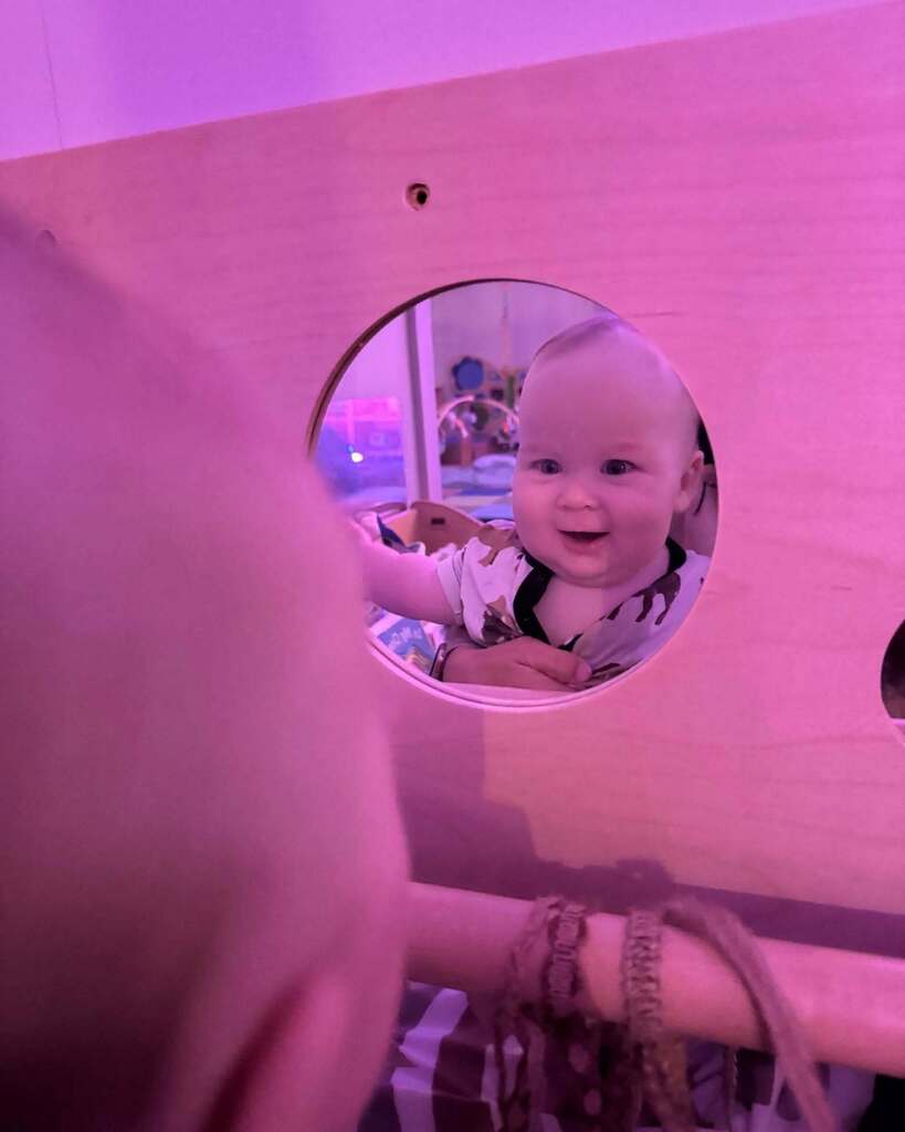 A baby in a crib looks at the camera with a smiling face through a round mirror mounted on the crib wall.