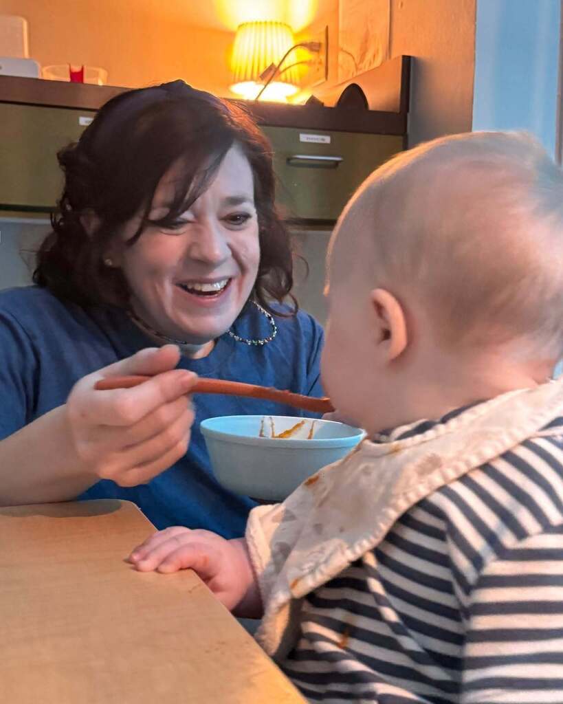 A woman holds a spoon in a bowl, feeding a baby sitting at a table.