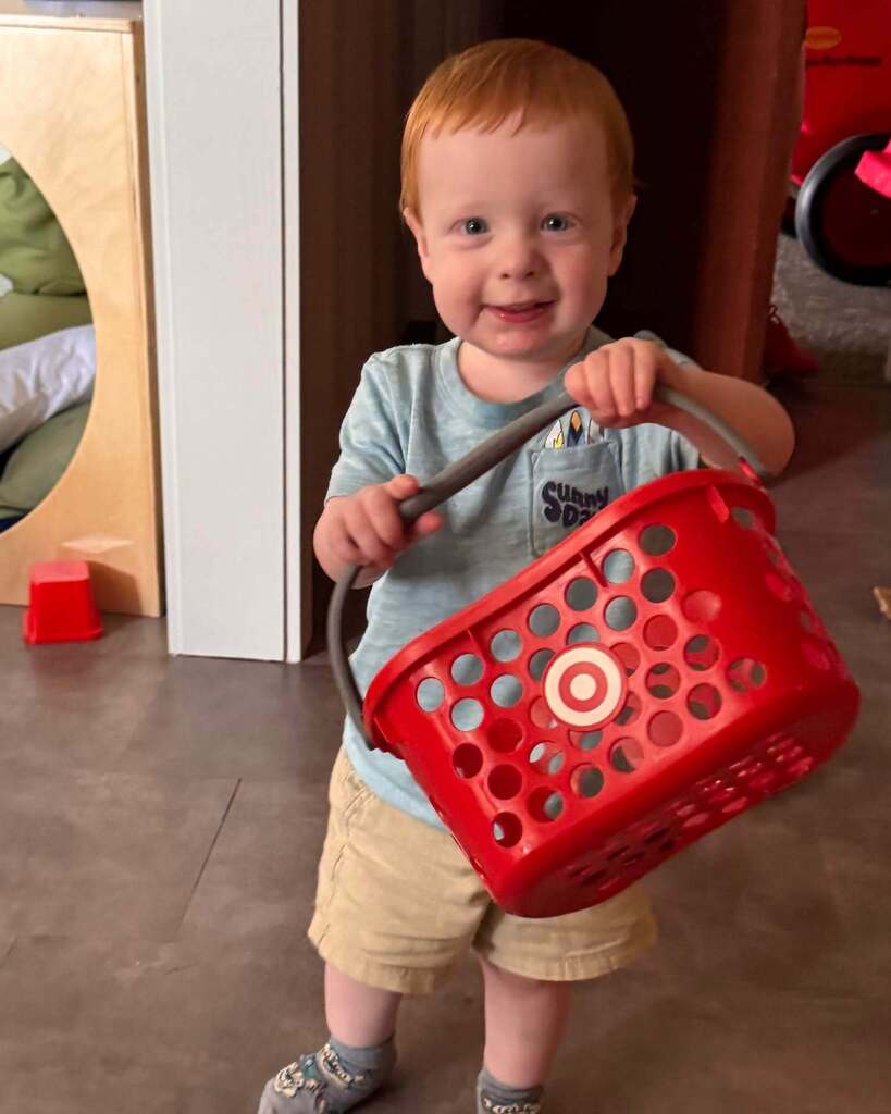 A young child, smiling, holding a red shopping basket. The room has light flooring and a door.