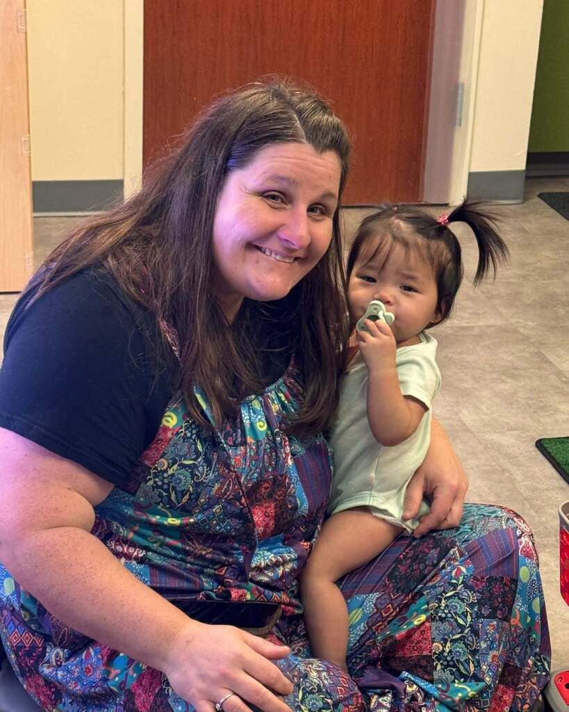 Woman wearing a colorful apron, seated on a floor, smiling and holding a baby on her lap.