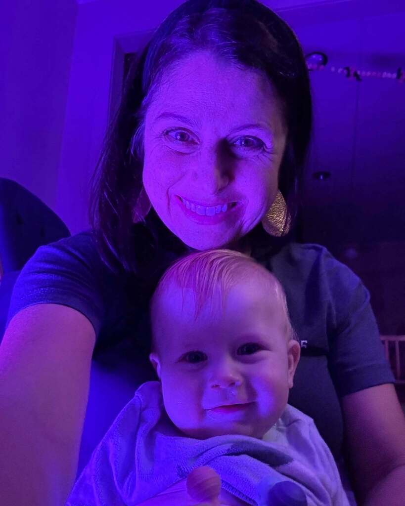 A smiling woman holding a smiling baby. Both have bright purple lighting behind them.
