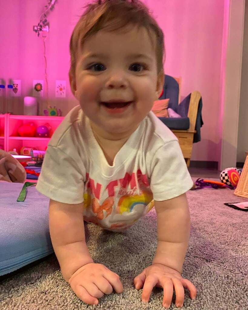 A baby in a white shirt crawls on the carpeted floor, smiling. A pink wall and toys are behind them.