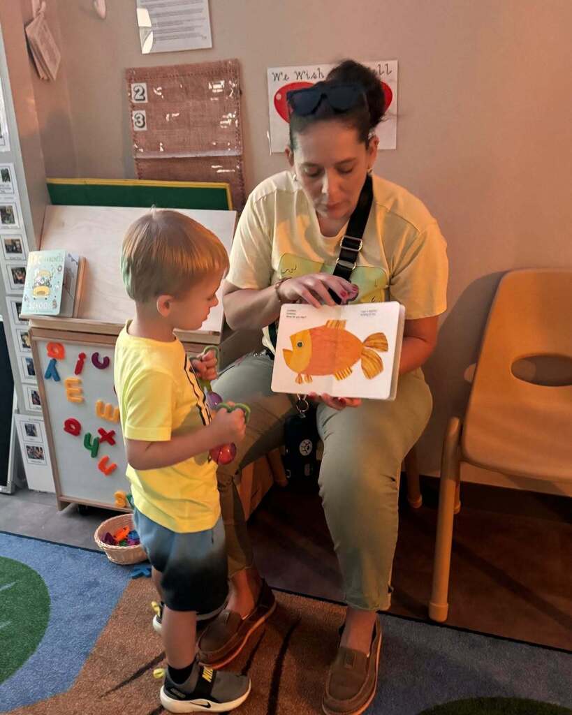 A woman in a yellow shirt is seated, reading a fish book to a toddler in a yellow shirt.
