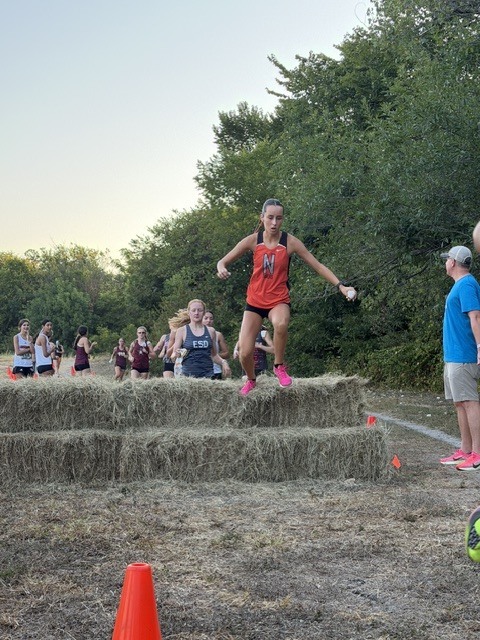 A runner in a red jersey jumps over hay bales in a field. People stand in the background.