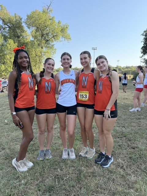 Five women in matching orange tops and black shorts. One wears a white shirt and number 153.