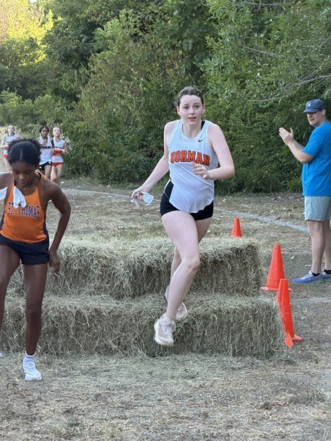 Two female runners in an outdoor cross-country event, with one jumping over hay bales and another running nearby. A coach watches from the side.