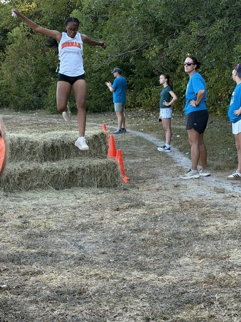 A female athlete in mid-jump over a straw hurdle, with a track and spectators in background.