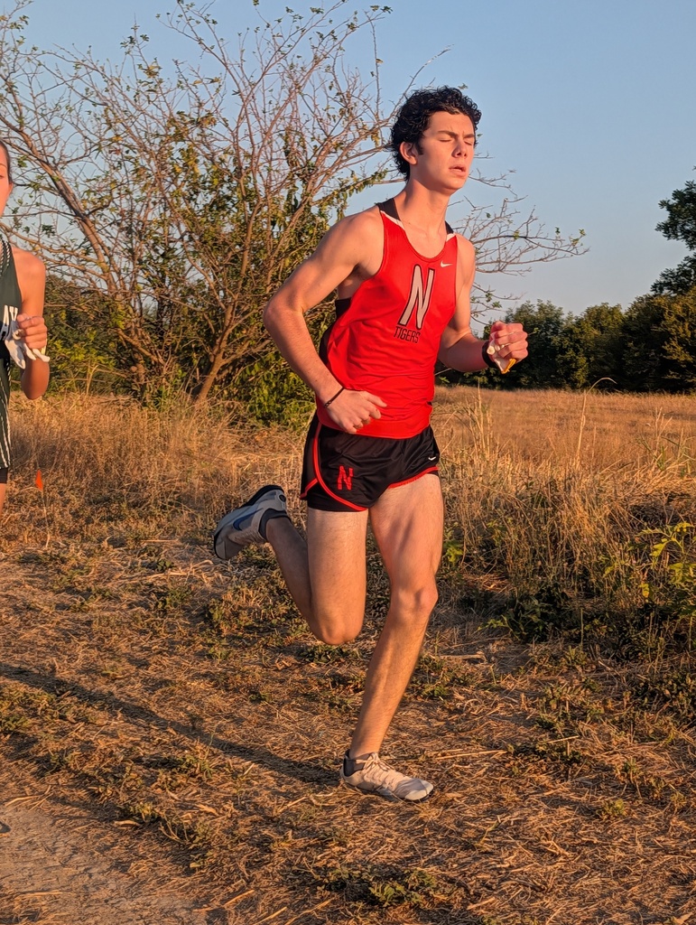 A runner in an orange tank top and black shorts runs on a dry grassy field.