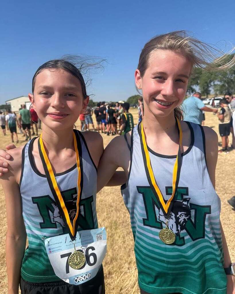 Two girls wearing green-and-white sports jerseys with medals around their necks, smiling. A field and people in the background.