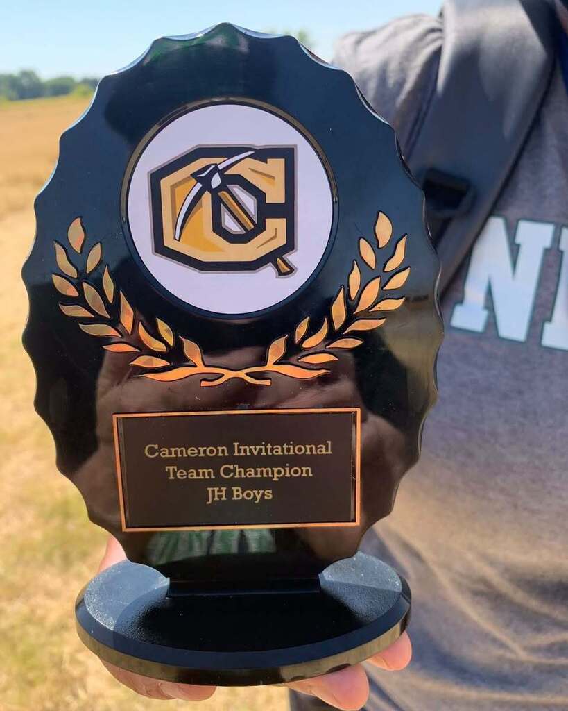 Person holds a trophy with a logo and the words "Cameron Invitational Team Champion Jr Boys." They are wearing a gray shirt.