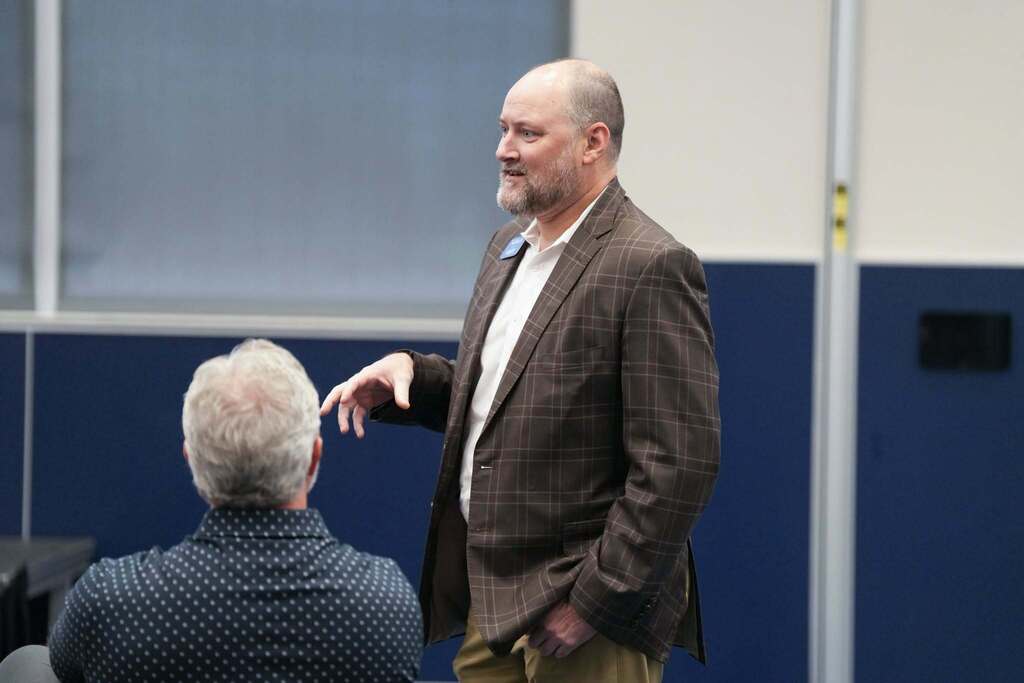 A man in a brown suit stands and speaks to another man with white hair seated in front of him in an indoor room.