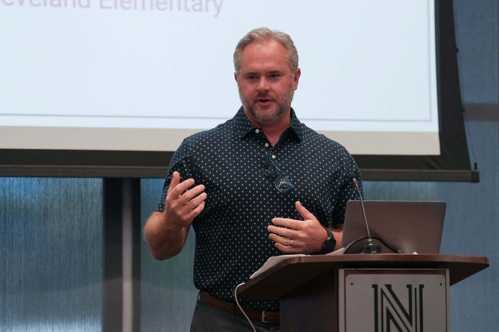 A man speaking at a podium with a laptop, gesturing with hands, and a projection screen behind.