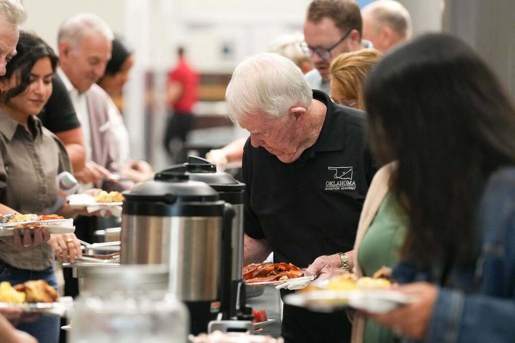 People gather at a buffet table, selecting various foods. One man wears a black shirt with an orange logo.
