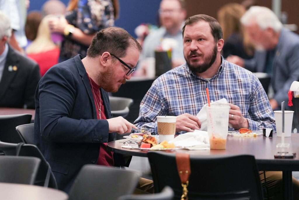Two men seated at a table with food, one in a suit and the other in a plaid shirt.