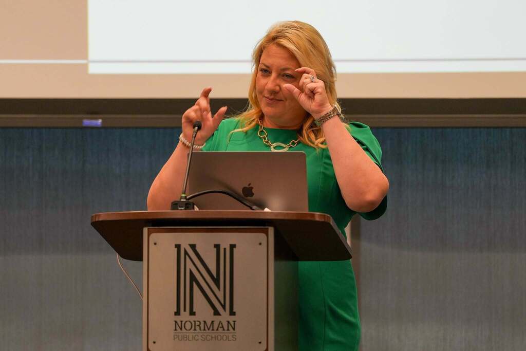A woman at a podium with a laptop, speaking to a room. Behind her is a screen, and the podium has an "N" logo.