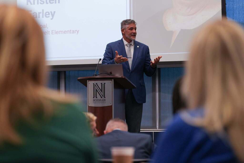 A man speaks at a podium with a microphone in front of a screen while several people listen.