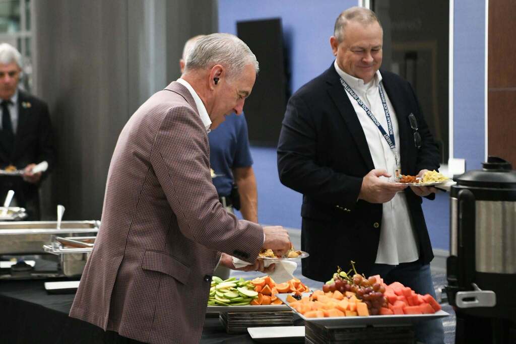 Two men select food from a buffet table with various dishes, one man holding a plate.