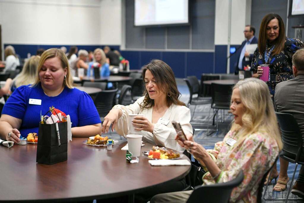 Four people sit at a round table in a conference hall. Three women face the camera, engaged in conversation.
