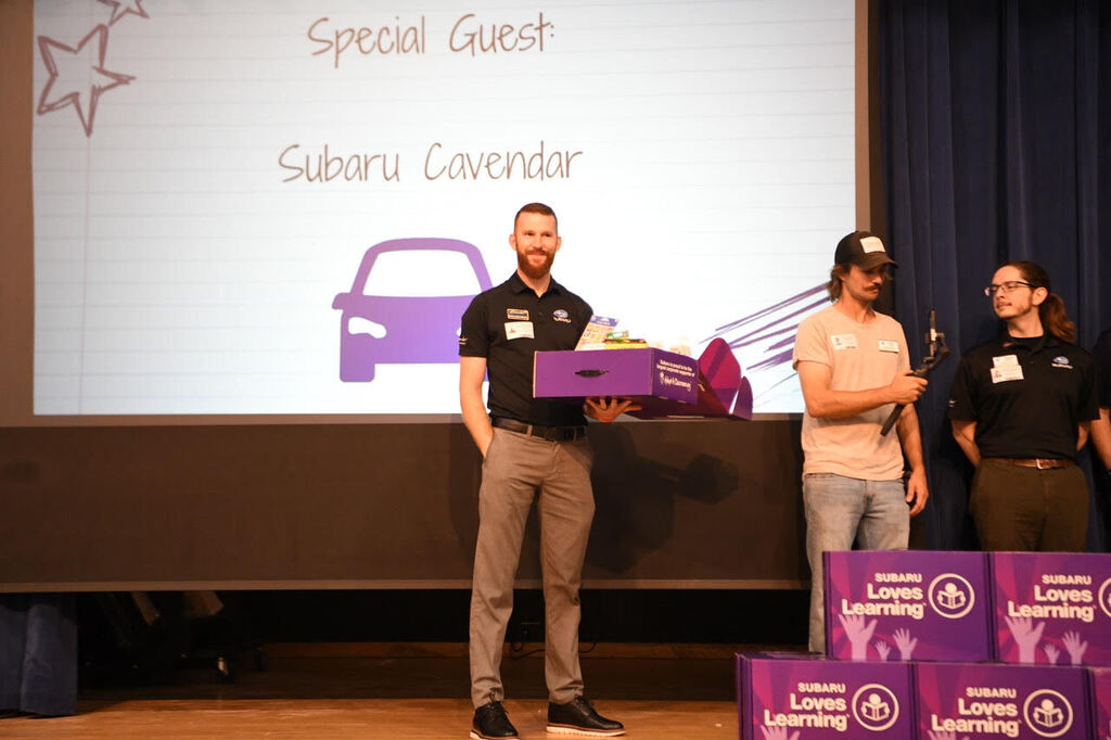 Three people on stage, one holding a purple box, another with a microphone. A projector screen behind them shows "Special Guest Subaru Cavendar" and a car image.
