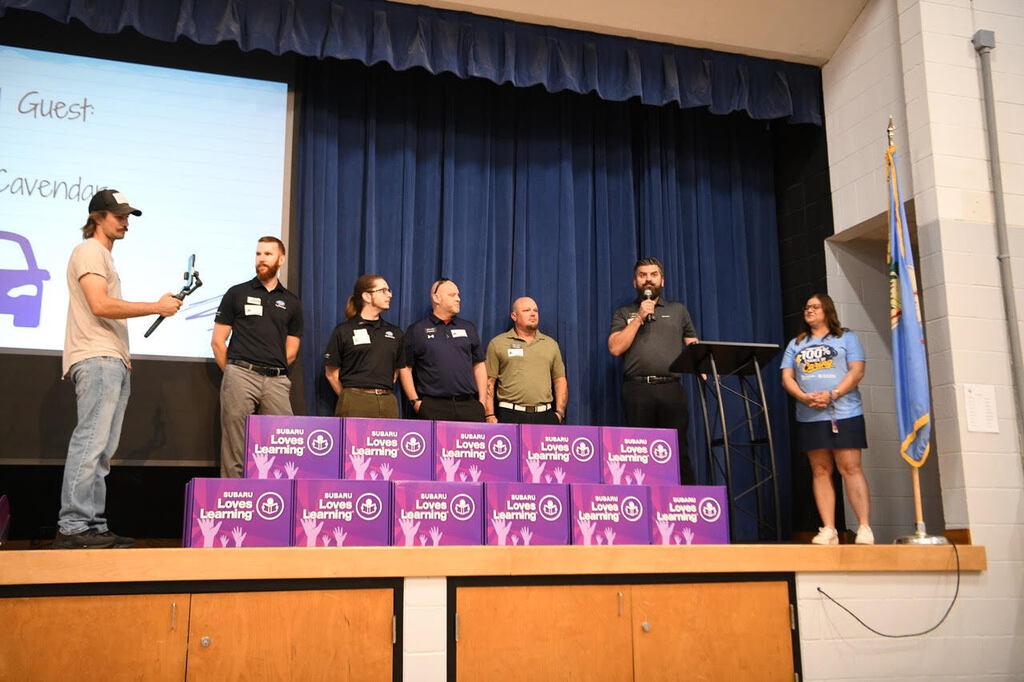 Group of people on stage in front of a screen and purple boxes. A flag is beside the stage.
