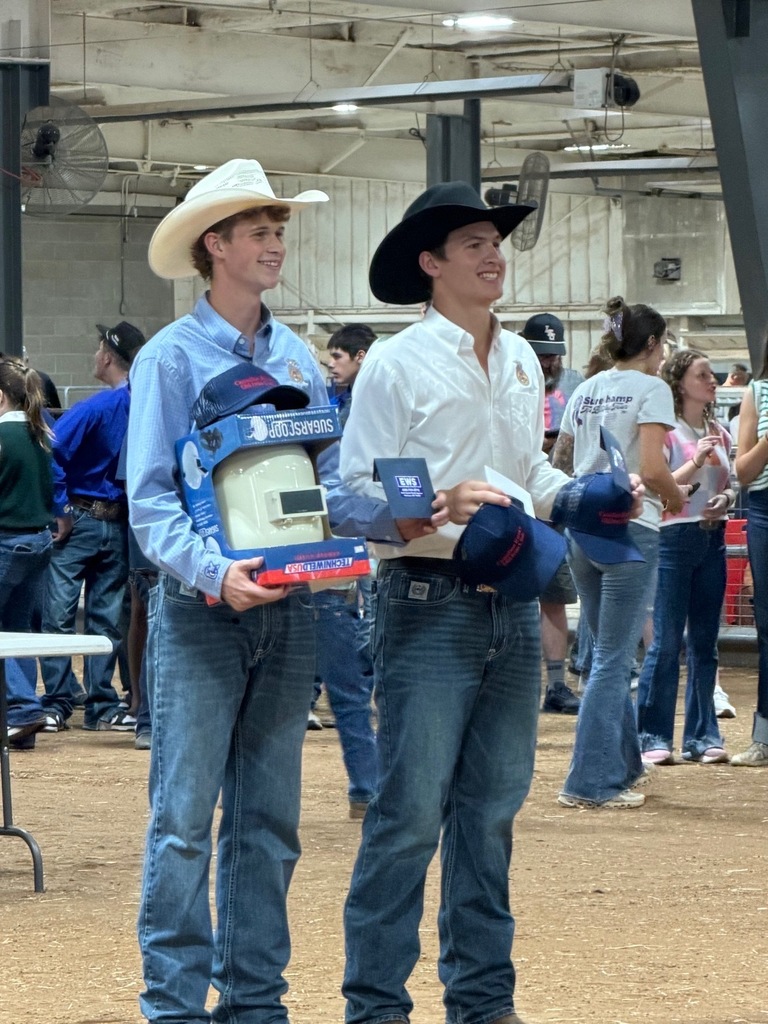 Two young men in cowboy hats and jeans stand next to each other, one holding a box and the other a hat.