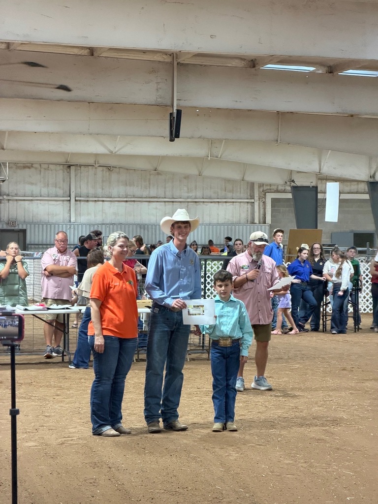 Three adults and a child stand in a dirt area with a crowd of people. One person holds a paper.