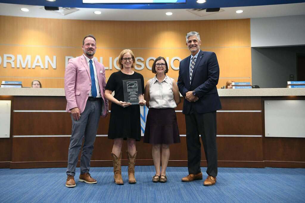 Four people stand together in front of a desk. The woman in the middle holds a trophy.