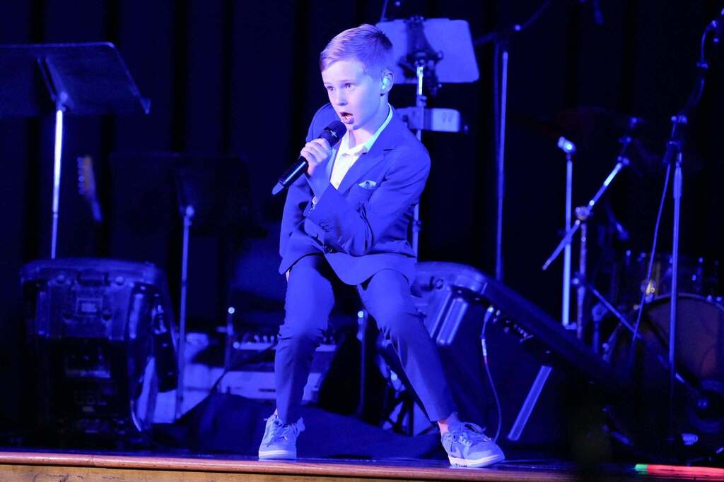 A young person in a blue suit on stage, holding a microphone, with speakers and instruments in the background.