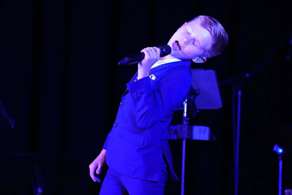 A young person in a blue suit sings into a microphone on a stage with blue lighting.