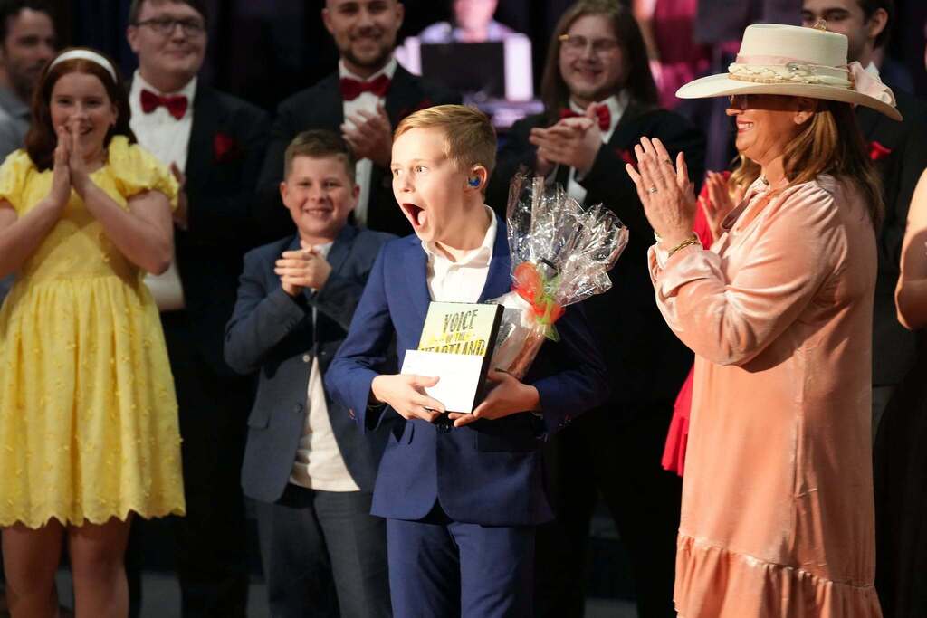 A boy in a blue suit holds a trophy, standing next to a woman in a pink dress. Behind them, several people watch, including a girl in a yellow dress and a man in a suit.