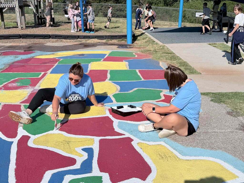 Two people in blue shirts are painting a colorful map of the US on the ground.