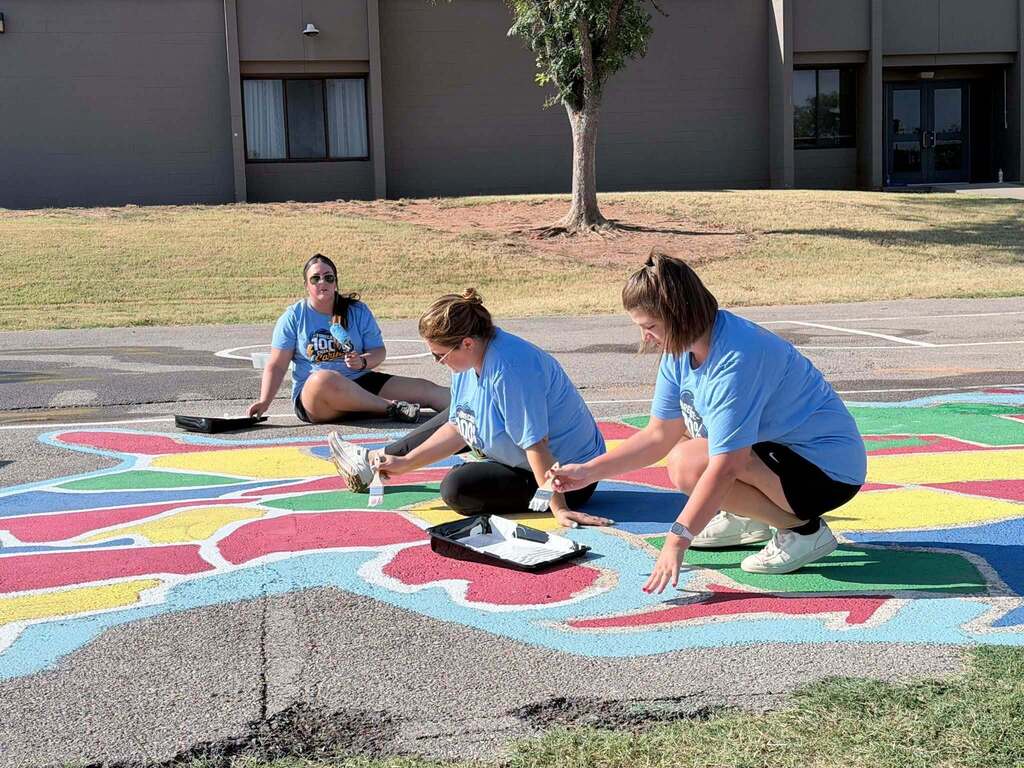 Three people in blue shirts and shorts kneeling to paint a multicolored flower in an open area.