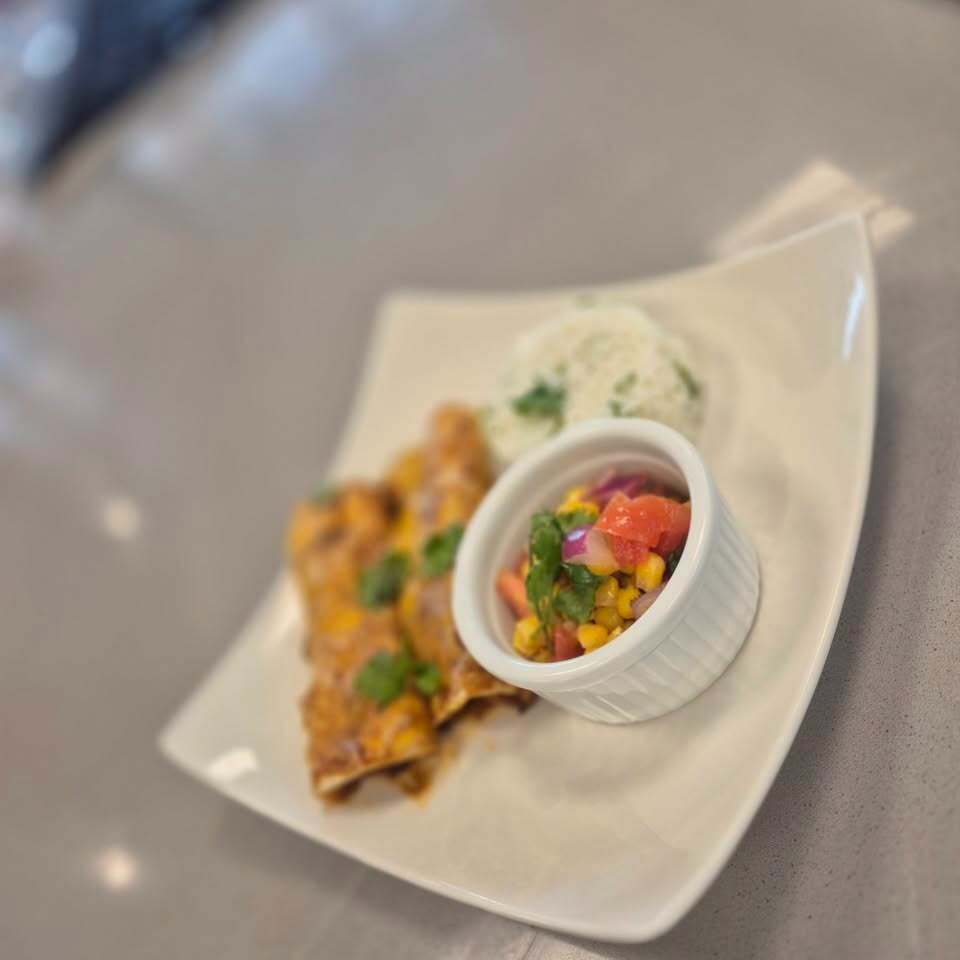 Square plate with two enchiladas and a white bowl with tomato and corn salsa on a gray table.
