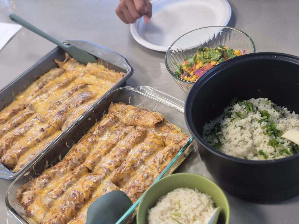 Trays of enchiladas and bowls of rice and salad on a table. An open container with a spoon in the center.