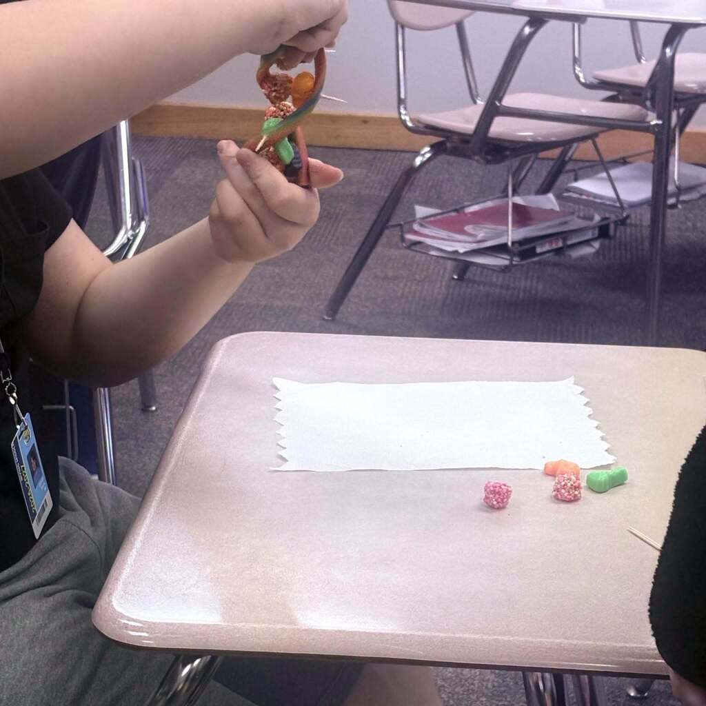 A person holds colorful beads on a desk with a white paper. Behind them, chairs and tables are arranged in rows.