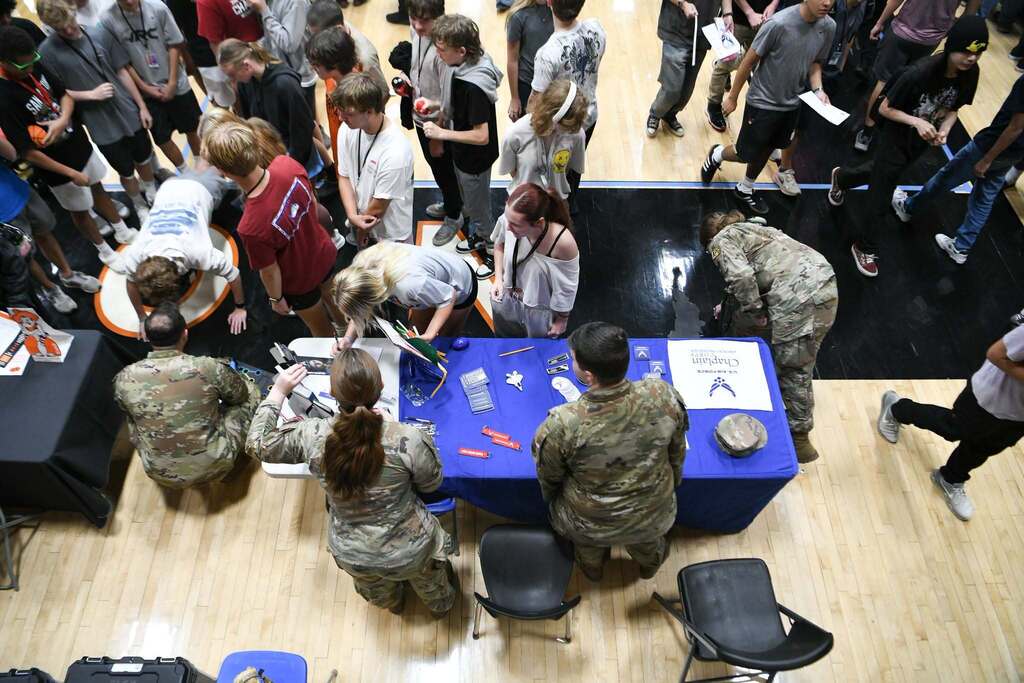 Several people in uniforms seated at a table with displays, surrounded by others looking on inside a gym.
