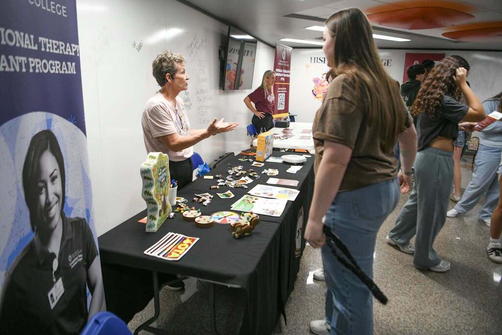 People in a hallway gather around a table with various items on display. One person gestures while speaking.