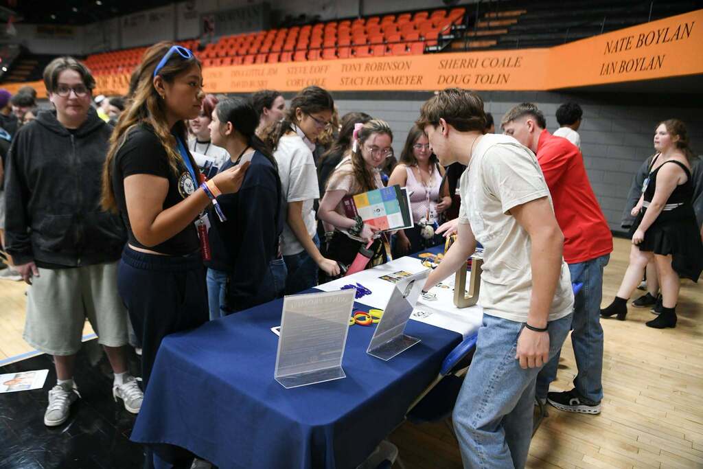 A group of people gather around a table with laptops, brochures, and a banner. Some wear casual clothing.