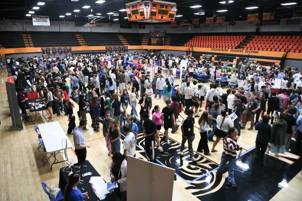 A large indoor area, possibly a gymnasium, is filled with a crowd of people. Many are standing and walking.