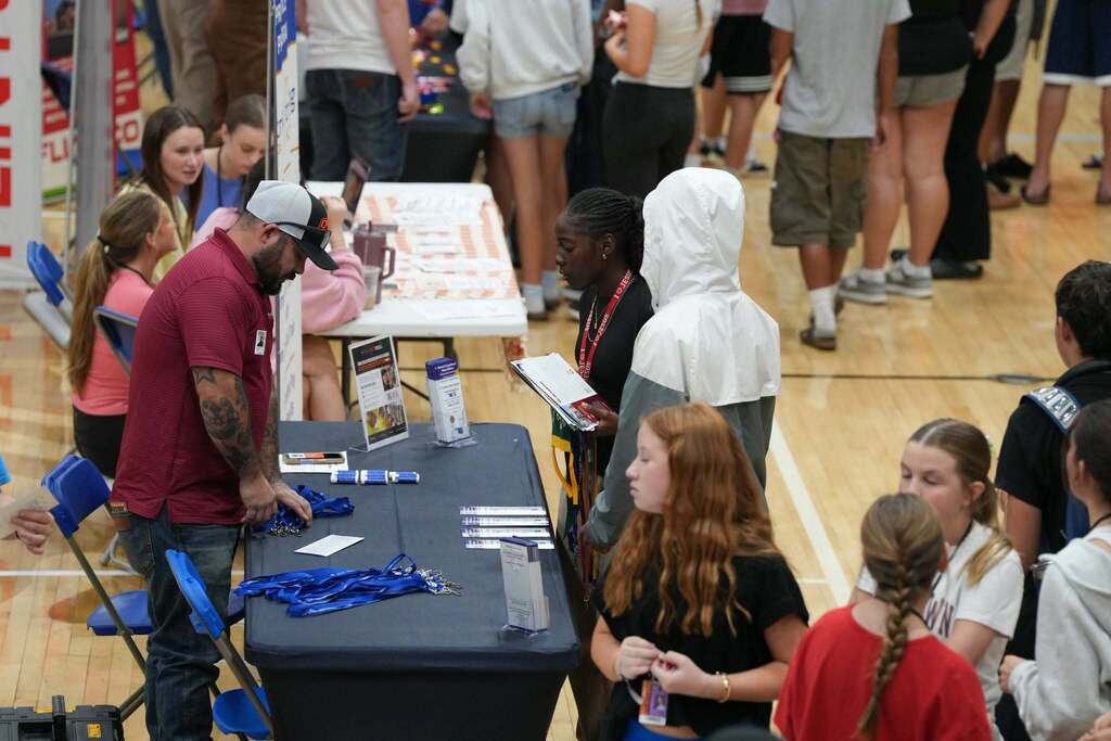 Group of individuals attending an indoor event. A person stands at a table with a table cloth, surrounded by a small crowd.