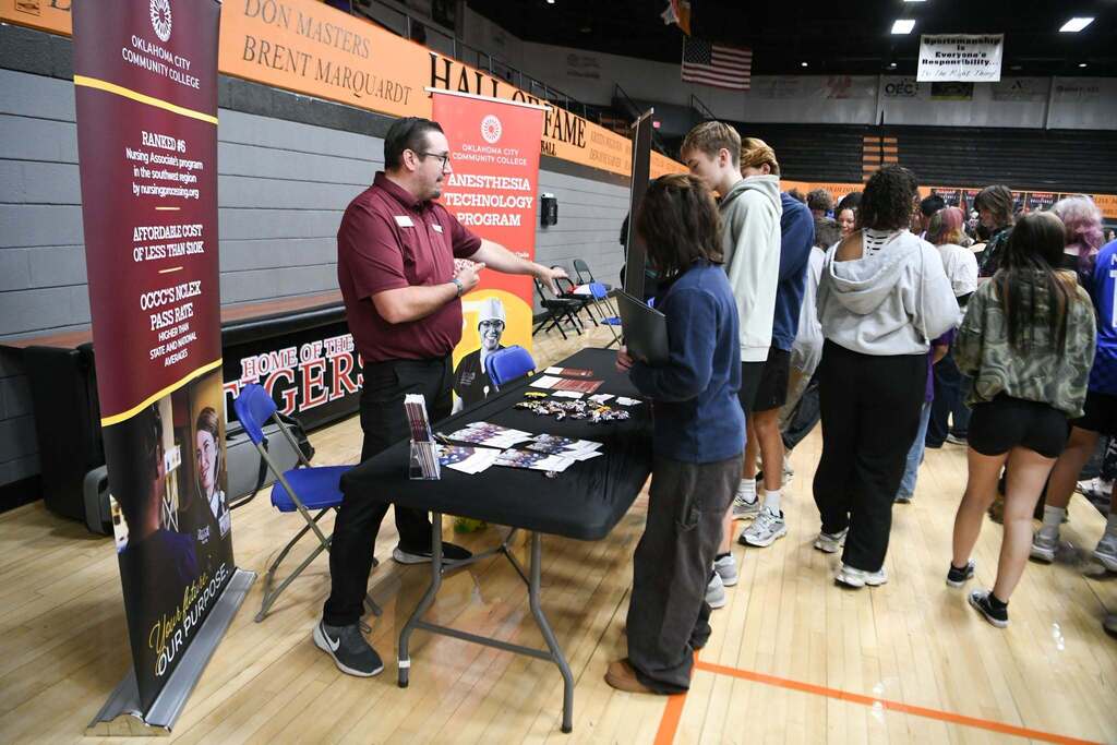 A man stands at a table in a gym, with a sign nearby, while several people gather behind him.