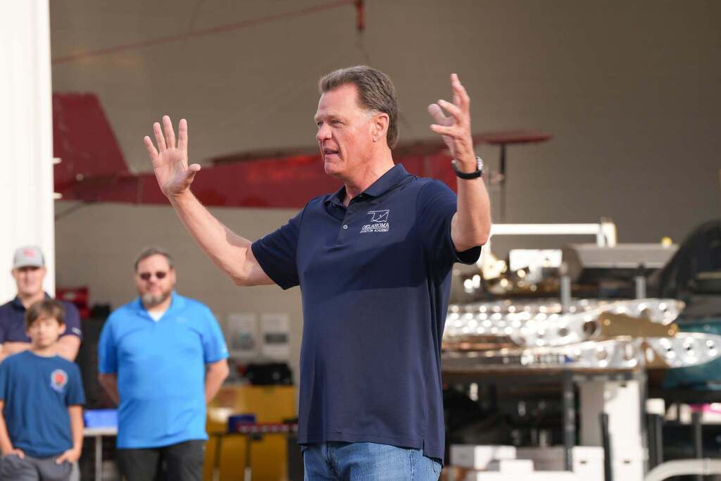A man speaking with his hands raised, wearing a navy blue polo, inside a hangar with a red plane.